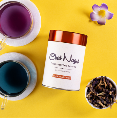 Blauer Tee von Chai Nagri in einer Packung mit zwei Tassen Tee und einer Blume auf gelbem Hintergrund.